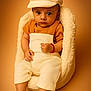 baby, infant, child, hat, overalls, orange_shirt, cream_color, fluffy_chair, soft_texture, studio_photo, portrait, sitting, cute, serious_expression, large_eyes, warm_tones, cozy, indoor, fashion, newborn