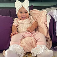 Aminah Thorton joined the competition — help win amazing prizes! baby, infant, smile, pink_clothing, white_bow, socks, blanket, pillow, bed, indoor, face, person, cute, happy, soft_toys, headwear, child, cozy, comfort, pacifier