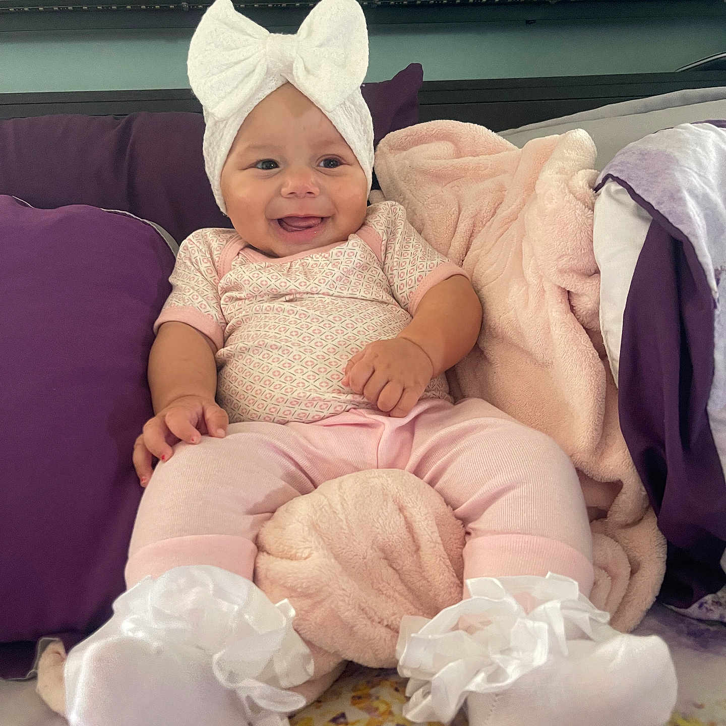 Aminah Thorton joined the competition — help win amazing prizes! baby, infant, smile, pink_clothing, white_bow, socks, blanket, pillow, bed, indoor, face, person, cute, happy, soft_toys, headwear, child, cozy, comfort, pacifier