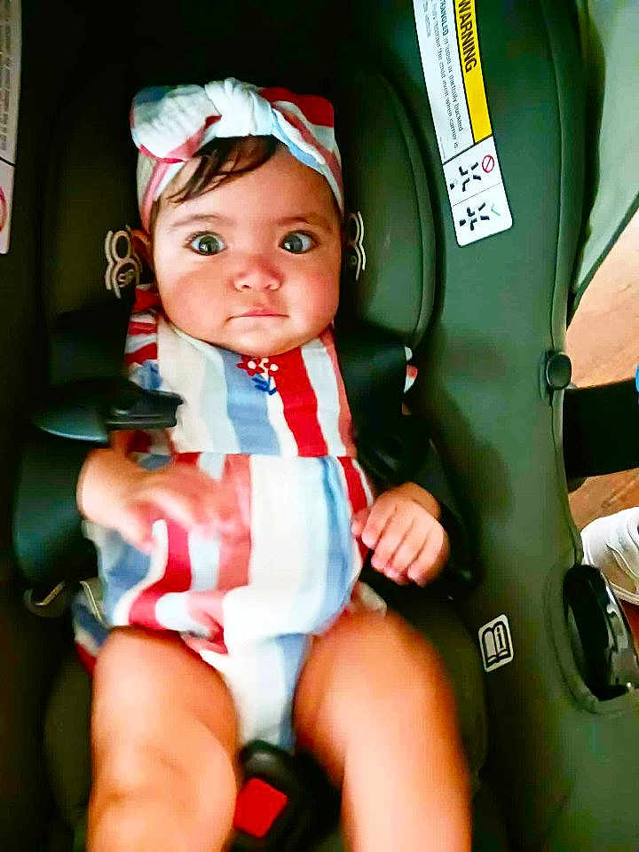 Reyna is registered to the contest to win money with this photo: baby, child, car_seat, striped_clothing, headband, wide_eyes, curious, sitting, safety_harness, infant, indoors, person, cute, chubby_legs, small_hands, colorful, secure, seatbelt, portrait, young