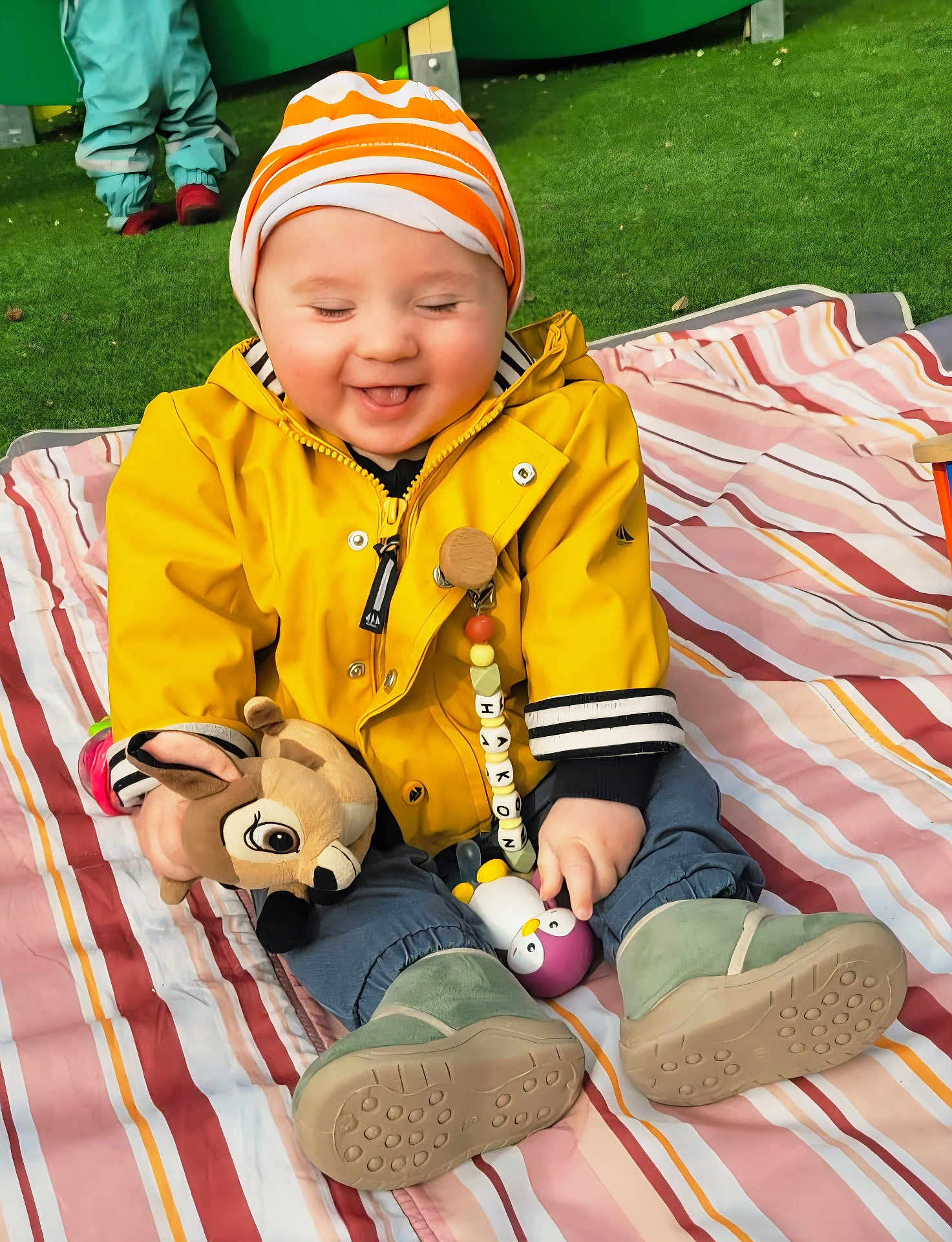 Hakon a rejoint le concours — aidez-le/la à gagner de superbes lots ! baby, infant, smiling, yellow_jacket, striped_hat, stuffed_toy, deer_toy, squeaky_toy, blanket, striped_blanket, grass, playground, boots, outdoor, seated, cute, happy, pacifier_clip, child, portrait