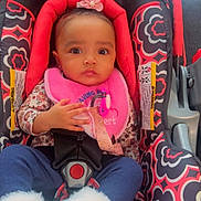 Ottilie is registered to the contest to win money with this photo: baby, child, car_seat, pink_bib, flower_headband, blue_pants, white_booties, patterned_fabric, indoor, sitting, infant, cute, person, young_child, seatbelt, clothing, portrait, looking_at_camera, adorable, closeup
