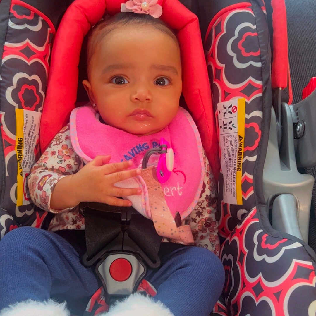 Ottilie is registered to the contest to win money with this photo: baby, child, car_seat, pink_bib, flower_headband, blue_pants, white_booties, patterned_fabric, indoor, sitting, infant, cute, person, young_child, seatbelt, clothing, portrait, looking_at_camera, adorable, closeup