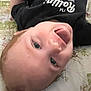 baby, smiling, tongue_out, black_shirt, text_on_shirt, floral_quilt, lying_down, child, happy, cute, infant, person, indoors, close_up, head, face, playful, skin, young_child, casual_clothing