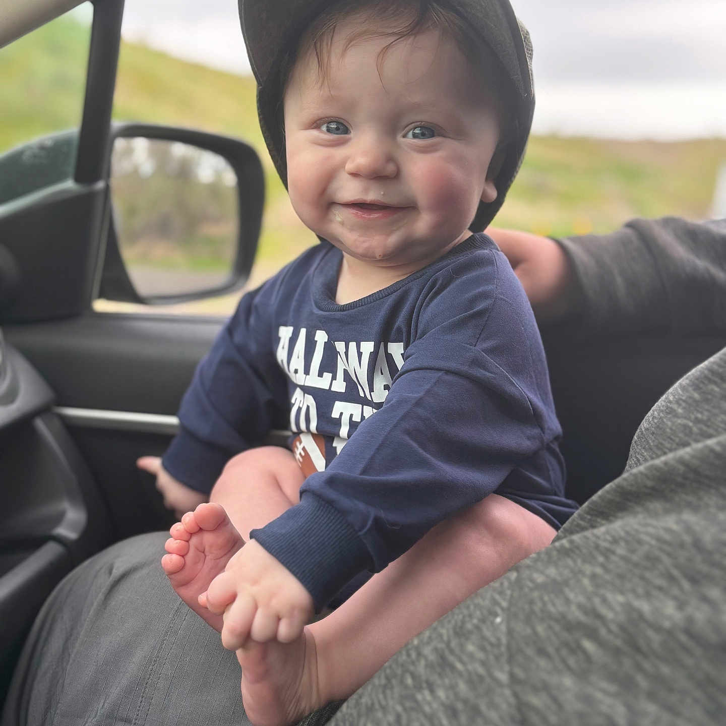 Fischer is registered to the contest to win money with this photo: adult, baby, blue_shirt, cap, car, car_interior, casual_clothing, child, face, foot, hand, happy, holding, indoors, person, seat, side_mirror, skin, smiling, window