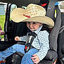 child, car_seat, cowboy_hat, blue_jeans, checkered_shirt, smiling, car_interior, seatbelt, young_child, cute, sitting, playful, vehicle, window, outdoor_view, door_open, seat, safety_harness, casual_clothing, portrait