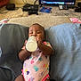 Miracle is registered to the contest to win money with this photo: baby, bottle, onesie, heart_pattern, cushion, blanket, living_room, television, gaming_console, carpet, floor, controller, child, indoors, person, milk, relaxing, cute, drink, home