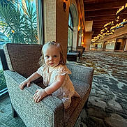 Amelia is registered to the contest to win money with this photo: toddler, child, armchair, indoor, hallway, window, plant, carpet, lighting, dress, lace, curious, furniture, person, portrait, blue_eyes, natural_light, architecture, decor, expression