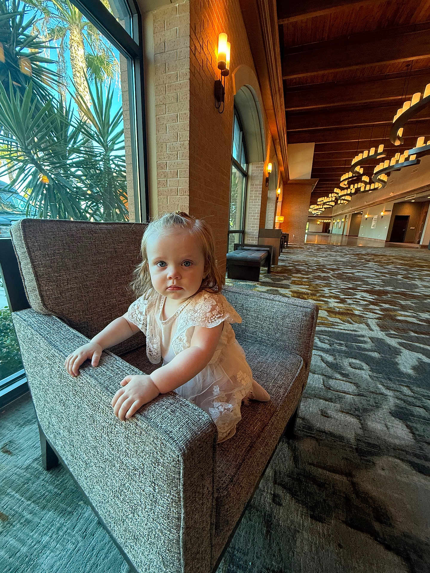 Amelia is registered to the contest to win money with this photo: toddler, child, armchair, indoor, hallway, window, plant, carpet, lighting, dress, lace, curious, furniture, person, portrait, blue_eyes, natural_light, architecture, decor, expression