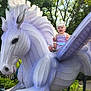 Nova is registered to the contest to win money with this photo: toddler, child, sculpture, winged_horse, pegasus, outdoor, garden, greenery, trees, sunlight, happy, smiling, headband, purple_clothing, playful, statue, nature, daylight, leaves, plants