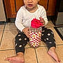 baby, barefoot, cabinet, checkered_pattern, child, curious_expression, curly_hair, floor, indoor, kitchen, long_sleeve_shirt, person, pink_hair_ties, sitting, smiling_flower, star_pattern_pants, tile_floor, toddler, water_bottle, wooden_cabinet