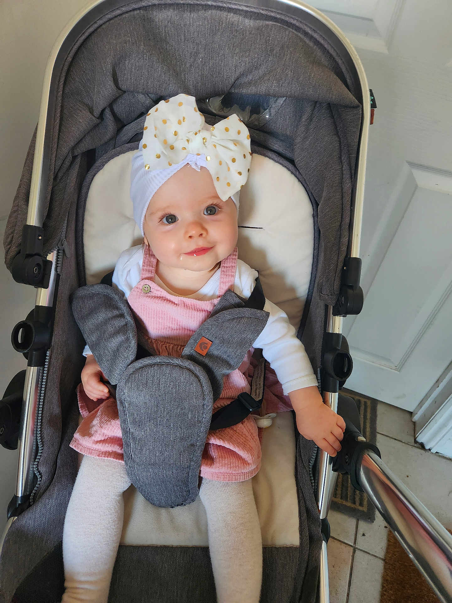 Zoé Lily is registered to the contest to win money with this photo: baby, infant, stroller, headband, bow, pink_dress, white_tights, smiling, drool, indoor, child, cute, seatbelt, metal_frame, floor_tiles, door, hand, clothing, person, portrait