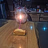 dog, birthday_candle, cake, table, wooden_table, kitchen, indoor, pet, curious, head_resting, candle_flame, lighting, household_items, counter, background_objects, domestic, animal, celebration, still_life, food