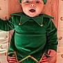 baby, infant, child, elf_costume, green_hat, striped_leggings, jingle_bells, costume, cute, surprised_expression, pink_cushion, lying_down, holiday, festive, clothing, indoors, portrait, smiling, face, person