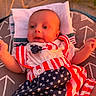 arm, baby, blue, bowtie, child, clothing, cushion, face, head, indoors, infant, laying, patriotic, pattern, red, smiling, stars, stripes, warm_light, white