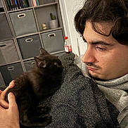 Titouni participe au concours pour gagner de l'argent avec cette photo : man, black_cat, kitten, indoor, sitting, holding, sweater, shelf, storage_bins, books, watch, facial_hair, curly_hair, closeup, pet, animal, home, cozy, caring, portrait
