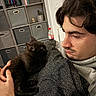 man, black_cat, kitten, indoor, sitting, holding, sweater, shelf, storage_bins, books, watch, facial_hair, curly_hair, closeup, pet, animal, home, cozy, caring, portrait