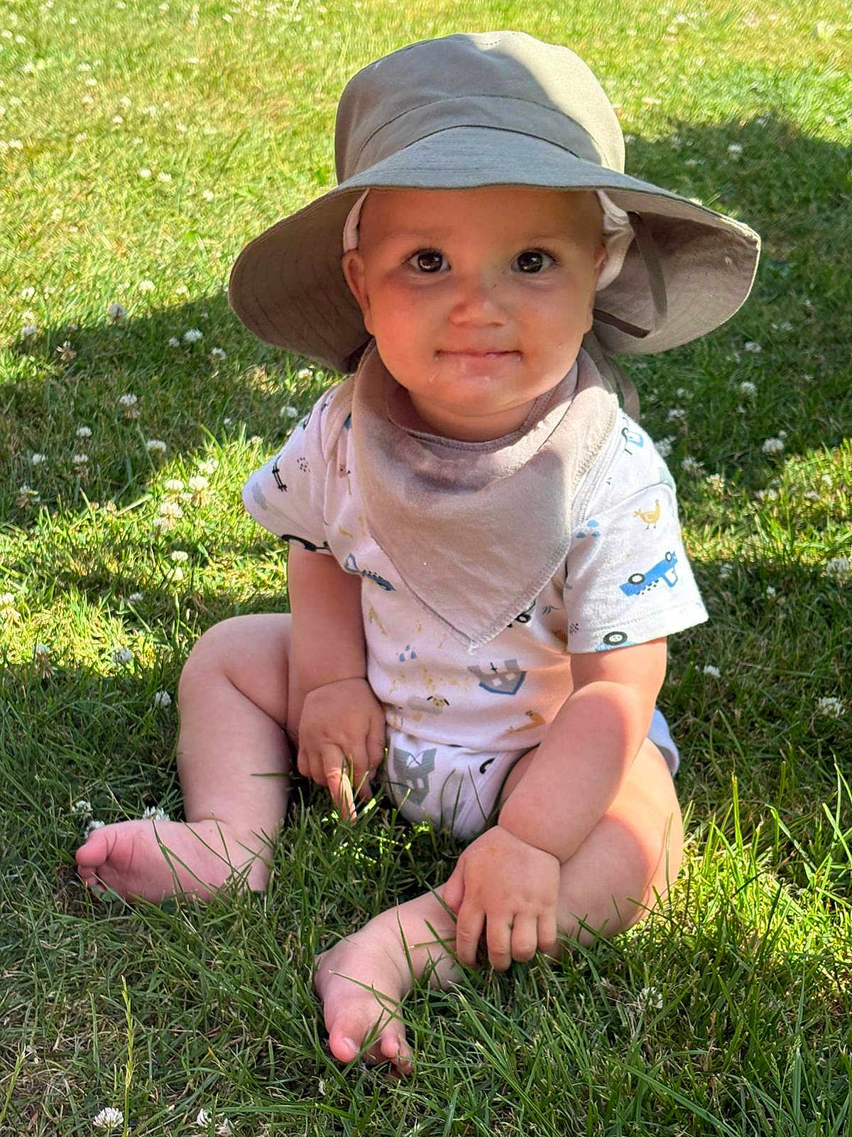 William is registered to the contest to win money with this photo: baby, child, grass, hat, sun_hat, outdoor, summer, greenery, sitting, cute, smile, bib, sunlight, nature, barefoot, infant, clothing, portrait, daylight, young