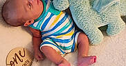 Asher is registered to the contest to win money with this photo: baby, infant, onesie, striped_clothing, stuffed_animal, plush_toy, blue_toy, blanket, soft_texture, wooden_sign, age_marker, newborn, cute, lying_down, indoors, child, person, feet, head, milestone