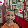 child, boy, smile, orange_shirt, indoor, diner, red_tiles, vintage, decor, counter, framed_photos, portrait, happy, casual_clothing, restaurant, background, young, person, retail, bright_lighting