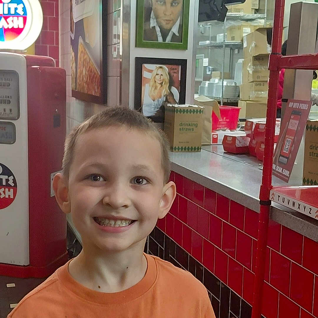 Carson joined the competition — help win amazing prizes! child, boy, smile, orange_shirt, indoor, diner, red_tiles, vintage, decor, counter, framed_photos, portrait, happy, casual_clothing, restaurant, background, young, person, retail, bright_lighting