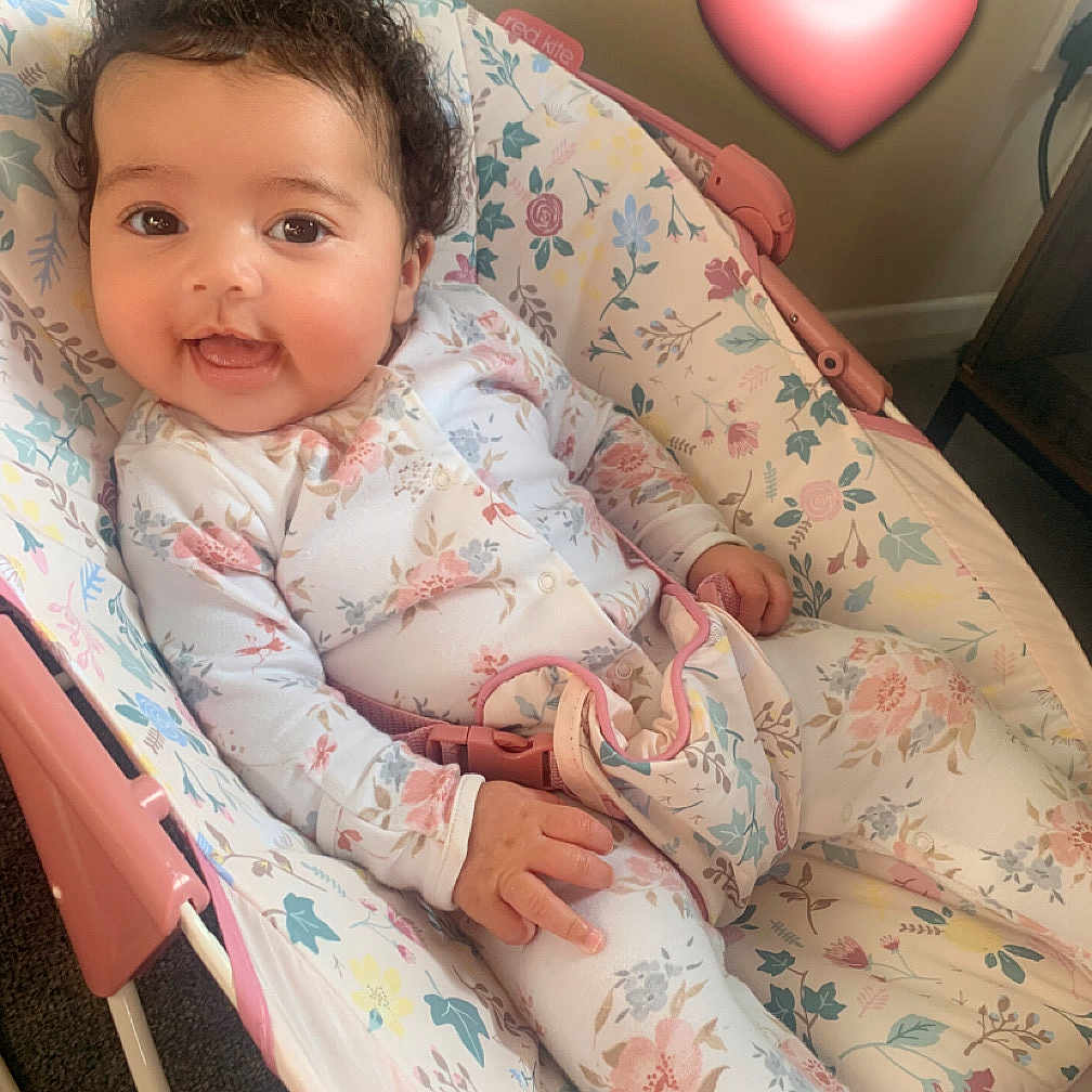 Cami is registered to the contest to win money with this photo: baby, baby_belt, baby_rocker, carpet, child, comfort, curly_hair, cute, floral_pattern, happy, heart_emoji, indoor, infant, onesie, person, pink, portrait, relaxed, seat, smiling
