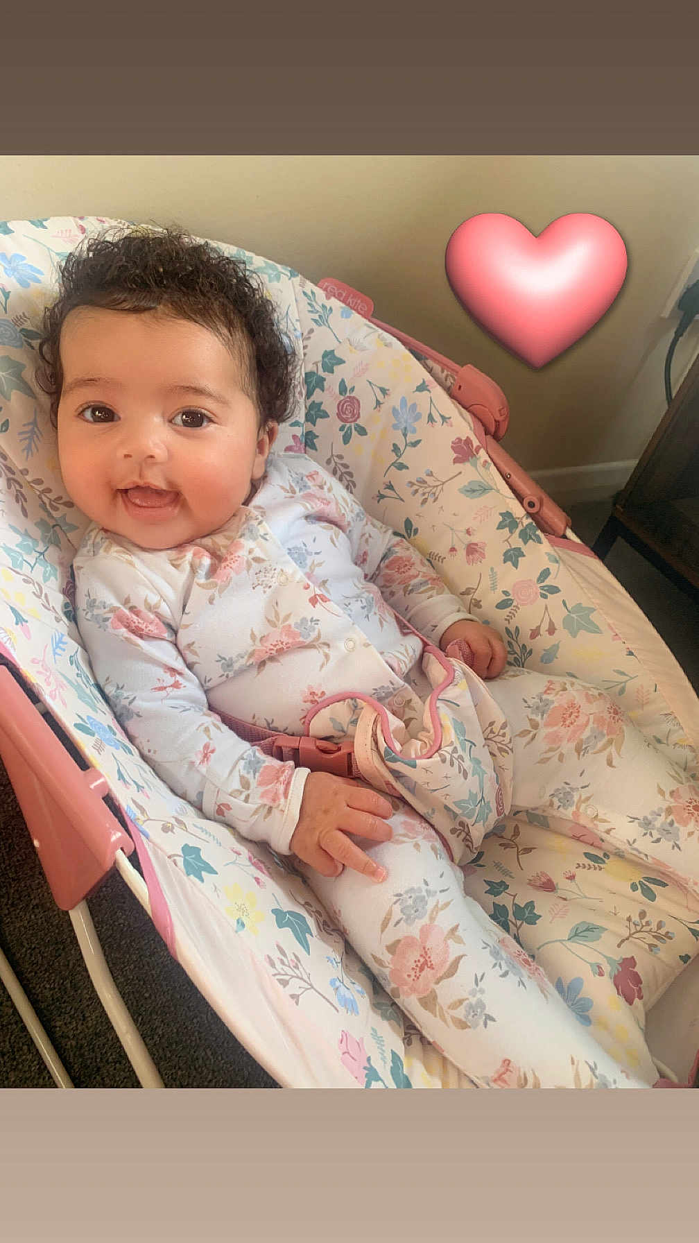 Cami is registered to the contest to win money with this photo: baby, infant, smiling, floral_pattern, onesie, baby_rocker, pink, heart_emoji, curly_hair, indoor, carpet, baby_belt, child, cute, happy, person, seat, comfort, relaxed, portrait