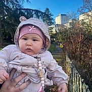 Leya a rejoint le concours — aidez-le/la à gagner de superbes lots ! baby, infant, child, outdoor, park, hat, hood, jacket, pacifier_clip, beaded_chain, hand, person, sky, trees, building, fence, pathway, grass, portrait, cute