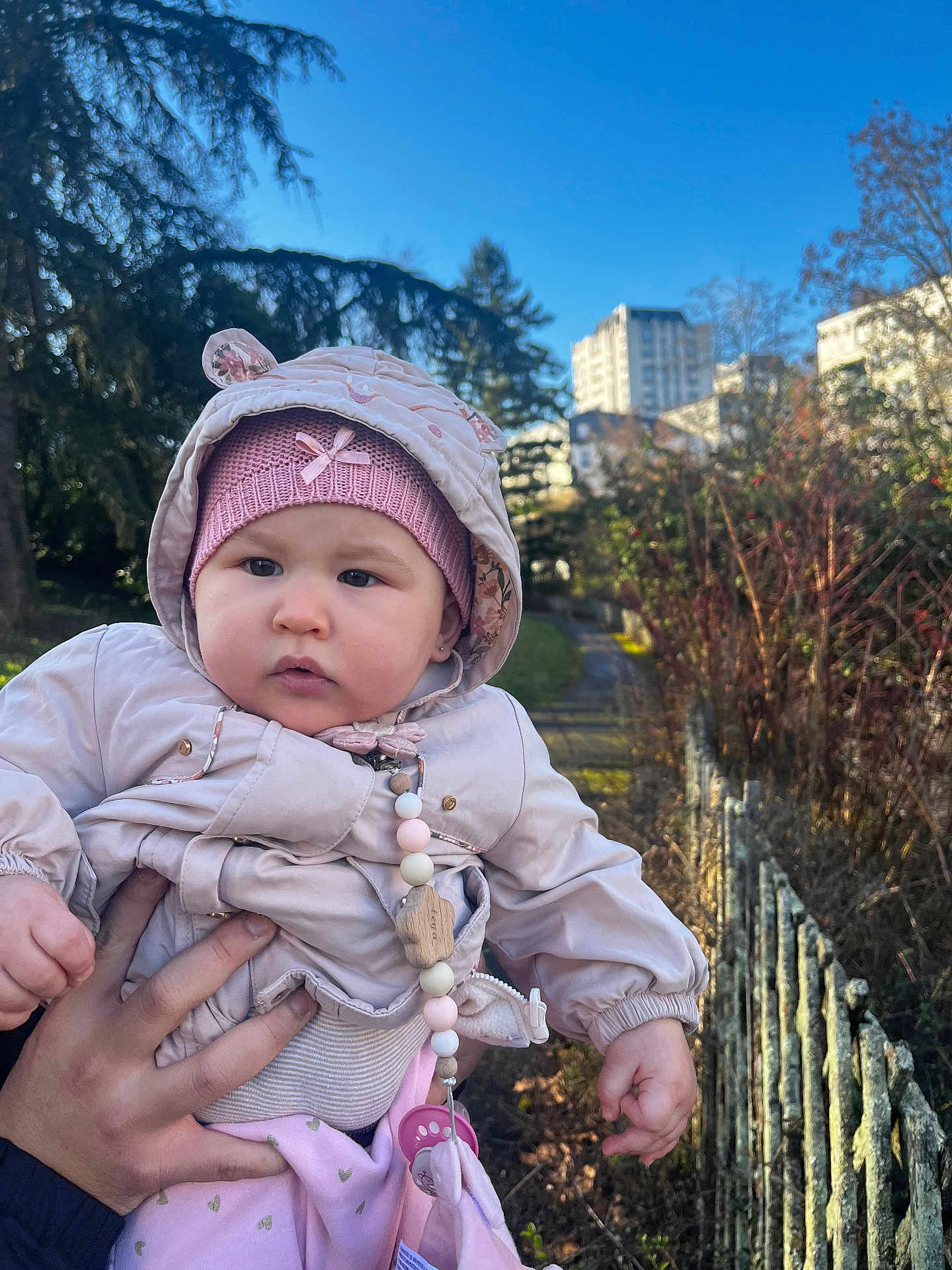 Leya a rejoint le concours — aidez-le/la à gagner de superbes lots ! baby, infant, child, outdoor, park, hat, hood, jacket, pacifier_clip, beaded_chain, hand, person, sky, trees, building, fence, pathway, grass, portrait, cute