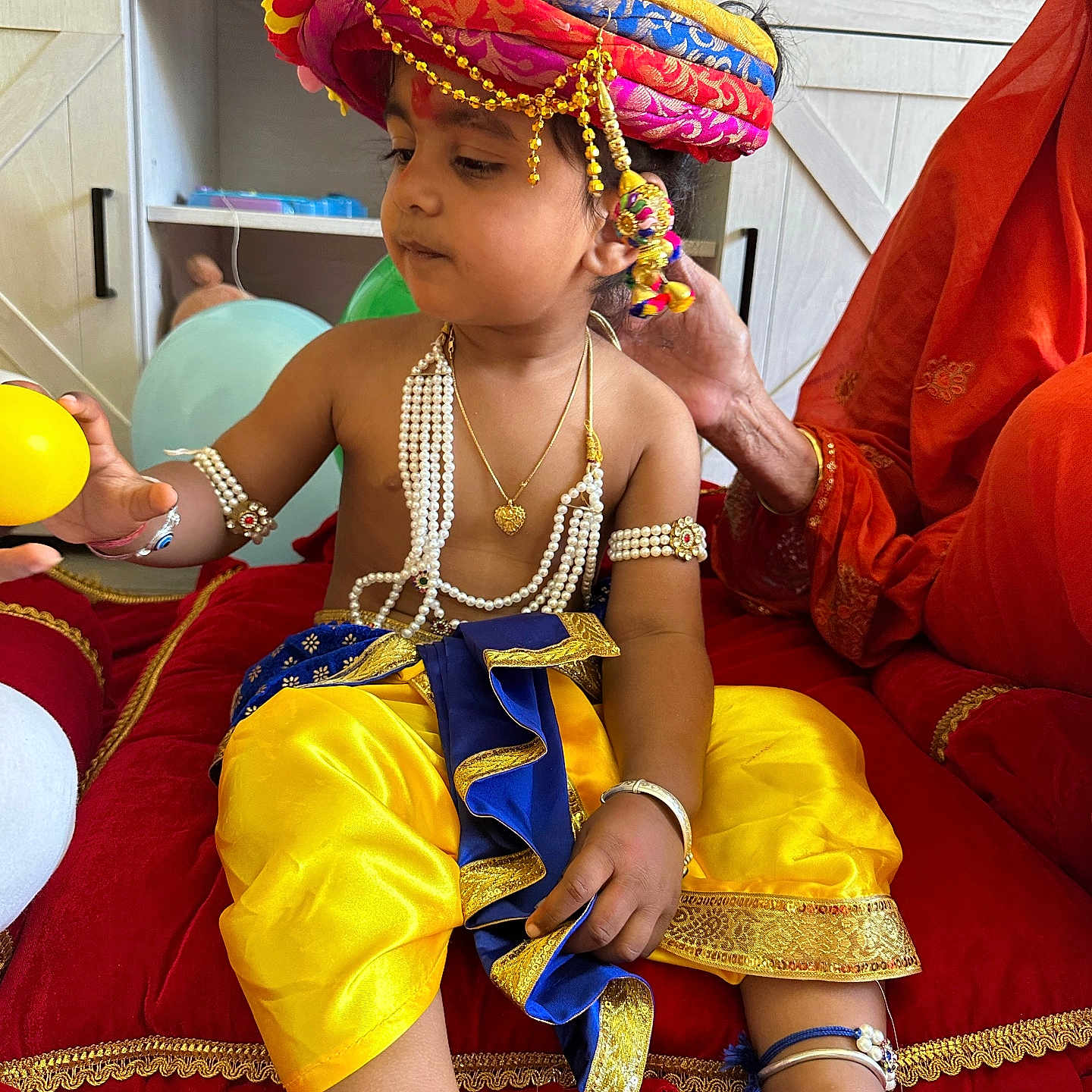Manas is registered to the contest to win money with this photo: balloon, blue_clothing, bracelet, child, curious, cushion, decor, furniture, hand, indoor, jewelry, pearl_necklace, person, red_fabric, sitting, television, toddler, traditional_clothing, turban, yellow_clothing