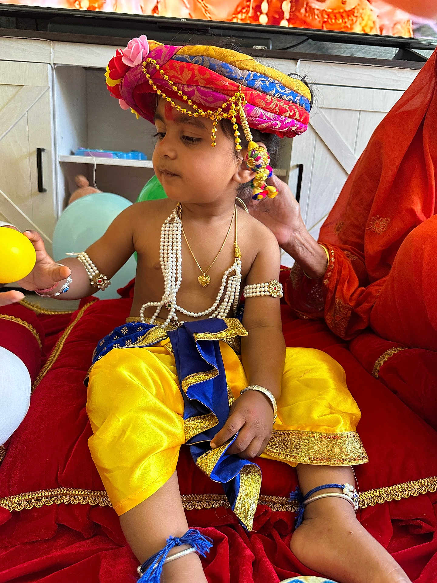 Manas is registered to the contest to win money with this photo: balloon, blue_clothing, bracelet, child, curious, cushion, decor, furniture, hand, indoor, jewelry, pearl_necklace, person, red_fabric, sitting, television, toddler, traditional_clothing, turban, yellow_clothing