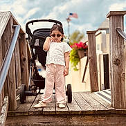 Siyara joined the competition — help win amazing prizes! child, toddler, sunglasses, white_top, pink_pants, shoes, stroller, wooden_bridge, handrail, american_flag, cloudy_sky, outdoor, flower_pot, wood, person, fashion, summer, cute, walking, portrait
