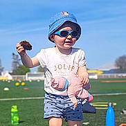 Clémence a rejoint le concours — aidez-le/la à gagner de superbes lots ! toddler, child, hat, sunglasses, doll, pinecone, shorts, shirt, sneakers, sports_field, green_grass, sunny, outdoor, toy, play, walking, blue_sky, water_bottle, fun, daytime