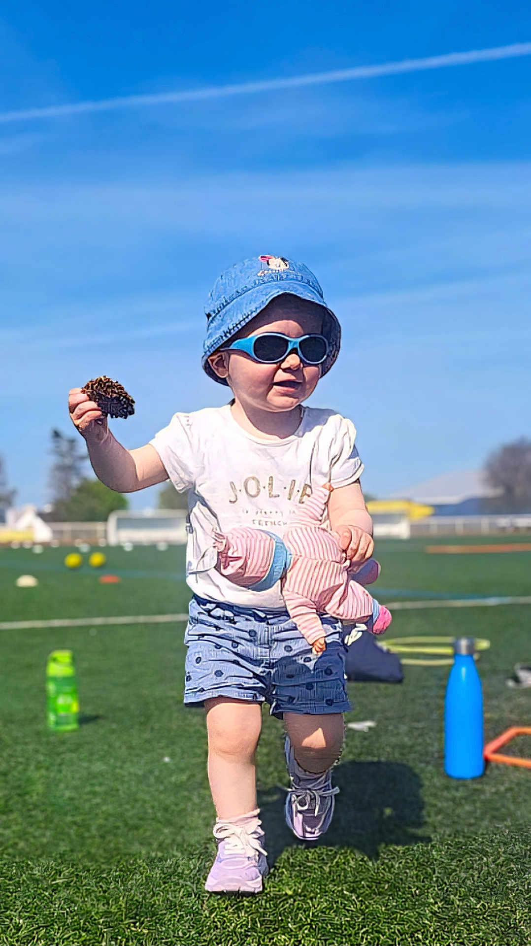 Clémence a rejoint le concours — aidez-le/la à gagner de superbes lots ! toddler, child, hat, sunglasses, doll, pinecone, shorts, shirt, sneakers, sports_field, green_grass, sunny, outdoor, toy, play, walking, blue_sky, water_bottle, fun, daytime