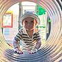 toddler, child, hat, striped_shirt, playground, tunnel, outdoor, hands, face, smile, person, window, building, daylight, fence, play_equipment, curious, cute, young_child, casual_clothing