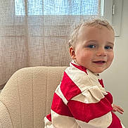 Noah participe au concours pour gagner de l'argent avec cette photo : toddler, child, smiling, red_and_white_striped_sweater, curly_hair, blue_eyes, beige_chair, indoor, curtain, natural_light, cozy, portrait, cute, happy, person, casual_clothing, seated, home_interior, young_child, soft_light