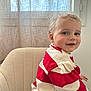 toddler, child, smiling, red_and_white_striped_sweater, curly_hair, blue_eyes, beige_chair, indoor, curtain, natural_light, cozy, portrait, cute, happy, person, casual_clothing, seated, home_interior, young_child, soft_light