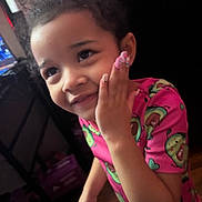 Iris is registered to the contest to win money with this photo: child, smiling, pink_shirt, avocado_pattern, hand_on_face, curly_hair, nails, indoor, portrait, person, happy, cute, young, girl, face, skin, wood_floor, furniture, dim_light, casual_clothing
