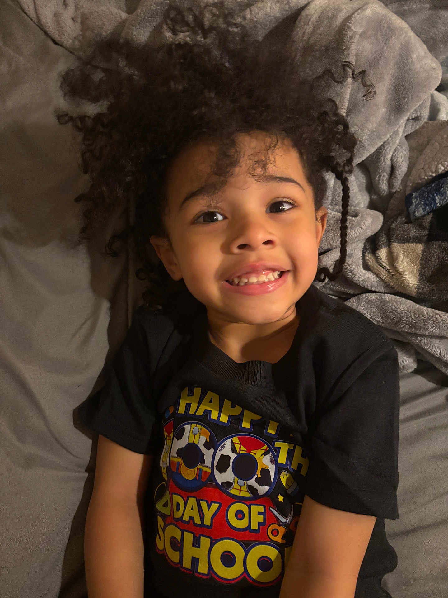 Iris joined the competition — help win amazing prizes! child, smiling, curly_hair, bed, blanket, bedding, tshirt, graphic_tshirt, portrait, face, eyes, teeth, happy, lying_down, indoor, young_child, person, closeup, black_clothing, cozy