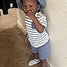 toddler, child, person, indoor, carpet, wall, clothing, hat, bucket_hat, shorts, striped_shirt, socks, pointing, standing, portrait, curious, young_child, home, casual, playful