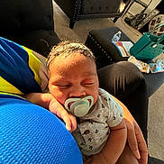 Adonis is registered to the contest to win money with this photo: baby, pacifier, sleeping, arm, blue_shirt, gray_onesie, anchor_pattern, hand, black_chair, ottoman, water_bottle, carpet, basket, paper, living_room, sunlight, indoor, person, comfort, resting
