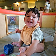 Lucas is registered to the contest to win money with this photo: baby, child, smiling, indoor, play_area, toys, blocks, bib, striped_shirt, happy, sitting, person, foot, hand, head, hair, face, background, adult, lighting
