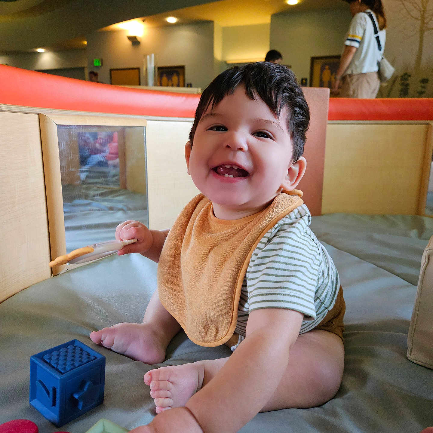 Lucas is registered to the contest to win money with this photo: adult, baby, background, bib, blocks, child, face, foot, hair, hand, happy, head, indoor, lighting, person, play_area, sitting, smiling, striped_shirt, toys