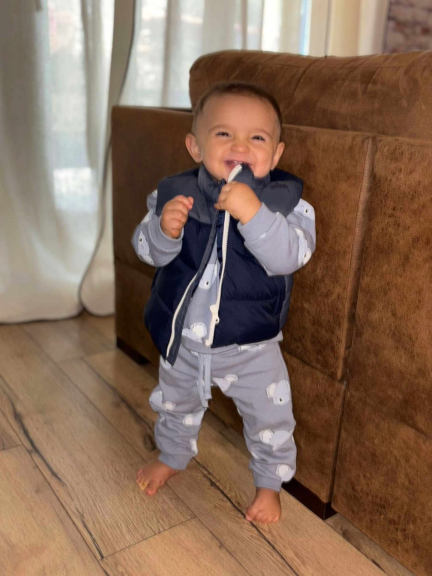 Ezio participe au concours pour gagner de l'argent avec cette photo : toddler, child, smiling, indoor, wooden_floor, brown_couch, navy_vest, grey_pajamas, koala_print, barefoot, curtains, playful, happy, standing, zipper, clothing, person, home_interior, cute, portrait