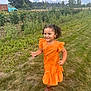 child, girl, orange_dress, boots, grass, flower_garden, sunflowers, vintage_truck, outdoor, smiling, curly_hair, nature, daylight, cloudy_sky, rural, greenery, playful, happy, portrait, casual