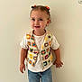 child, toddler, smile, curly_hair, hair_bows, colorful_vest, crocheted_vest, white_tshirt, blue_jeans, standing, indoor, plain_wall, happy, casual_clothing, person, cute, young_child, fashion, portrait, small_child