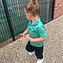 Aliya participe au concours pour gagner de l'argent avec cette photo : child, toddler, hair_bun, polo_shirt, teal_shirt, black_pants, white_sneakers, outdoor, side_view, curious, pebbled_pathway, metal_fence, brick_wall, casual_clothing, young_child, walking, daylight, person, motion, street
