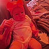 baby, infant, red_bow, onesie, hand, blanket, bed, fabric, indoor, person, child, holding_hand, cute, soft_lighting, relaxed, skin, headwear, comfort, warm_colors, seated