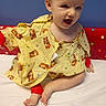 baby, child, hospital_gown, yellow_clothing, tiger_print, red_wristband, red_ankleband, polka_dot, bed, white_sheet, smiling, happy, sitting, infant, cute, face, indoor, person, young_child, playful