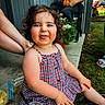 child, toddler, curly_hair, dress, outdoor, grass, porch, plastic_cup, snack_bag, person, sitting, happy, playful, summer, family, casual, daylight, cheerful, rosy_cheeks, tongue_out